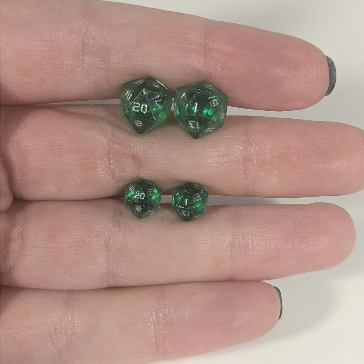 Two pairs of handmade d20 earrings with titanium hardware that are emerald green with silver numbers held between the fingers of a flat hand. The larger pair is between the pointer and middle finger, the smaller between the middle and ring finger. The earrings on the left of each pair centers the 20 face and the earrings on the right of each pair centers the 1 face.