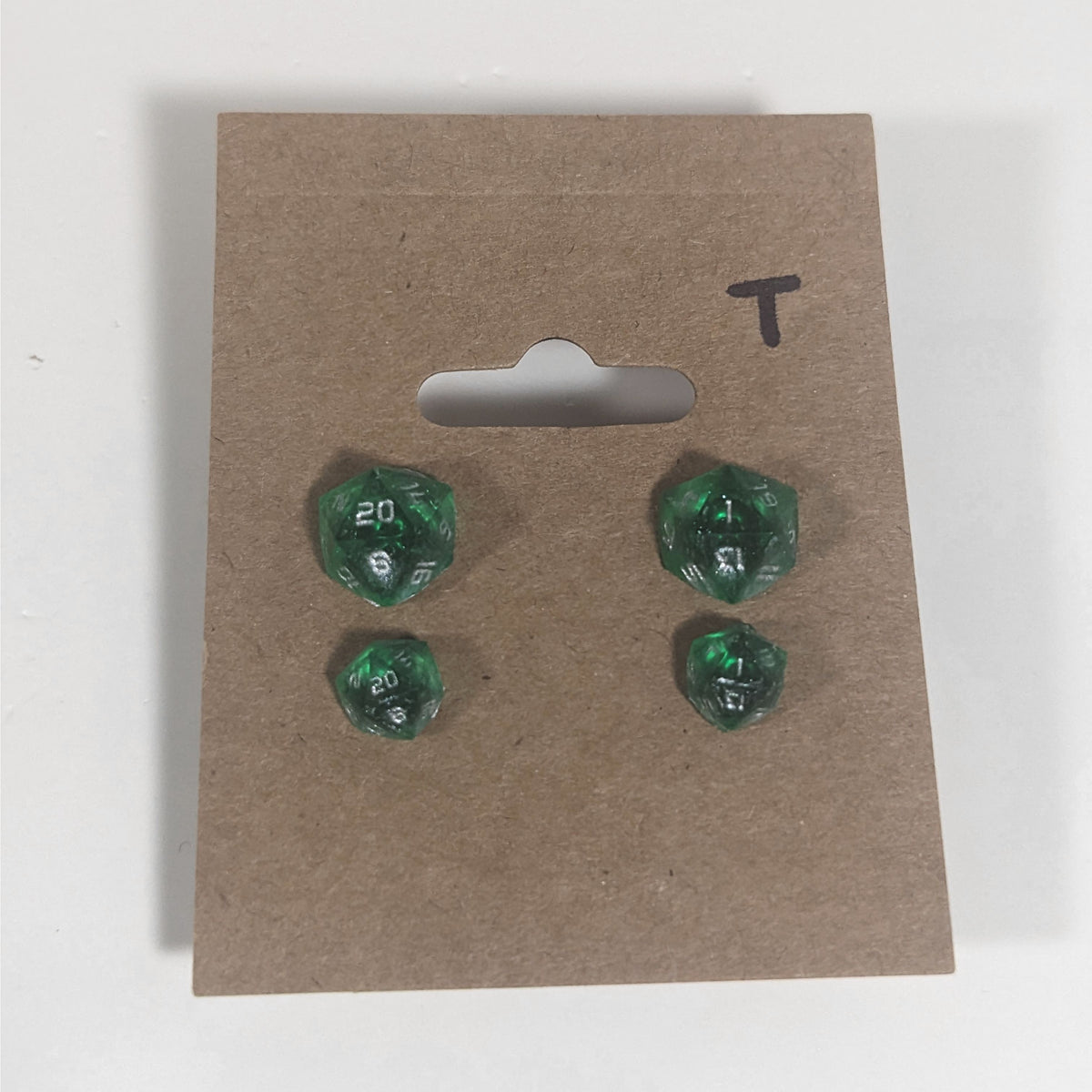 Two pairs of handmade d20 earrings with titanium hardware that are emerald green with silver numbers on a kraft brown earring card with a T in the top right corner. The earrings on the left of each pair centers the 20 face and the earrings on the right of each pair centers the 1 face.