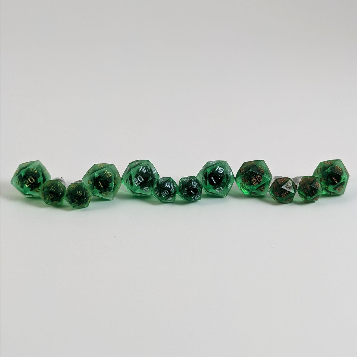 Six pairs of emerald green d20 stud earrings with titanium hardware on a white background. There are 3 large pairs and 3 small pairs, and one pair of each of those has gold numbers, silver numbers, and copper numbers.