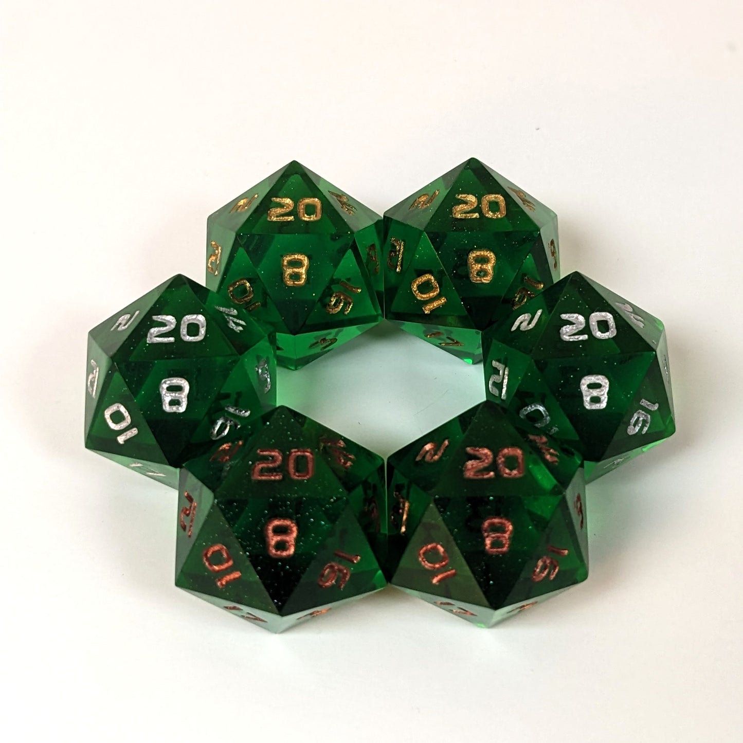 Three pairs of standard d20s are arranged in a hexagon on a white background. All of the handmade dice are emerald green. The top two d20s have gold numbers, the middle two have silver numbers, and the bottom two have copper numbers, showing how each number color affects the appearance of the hue of the emerald green dice.