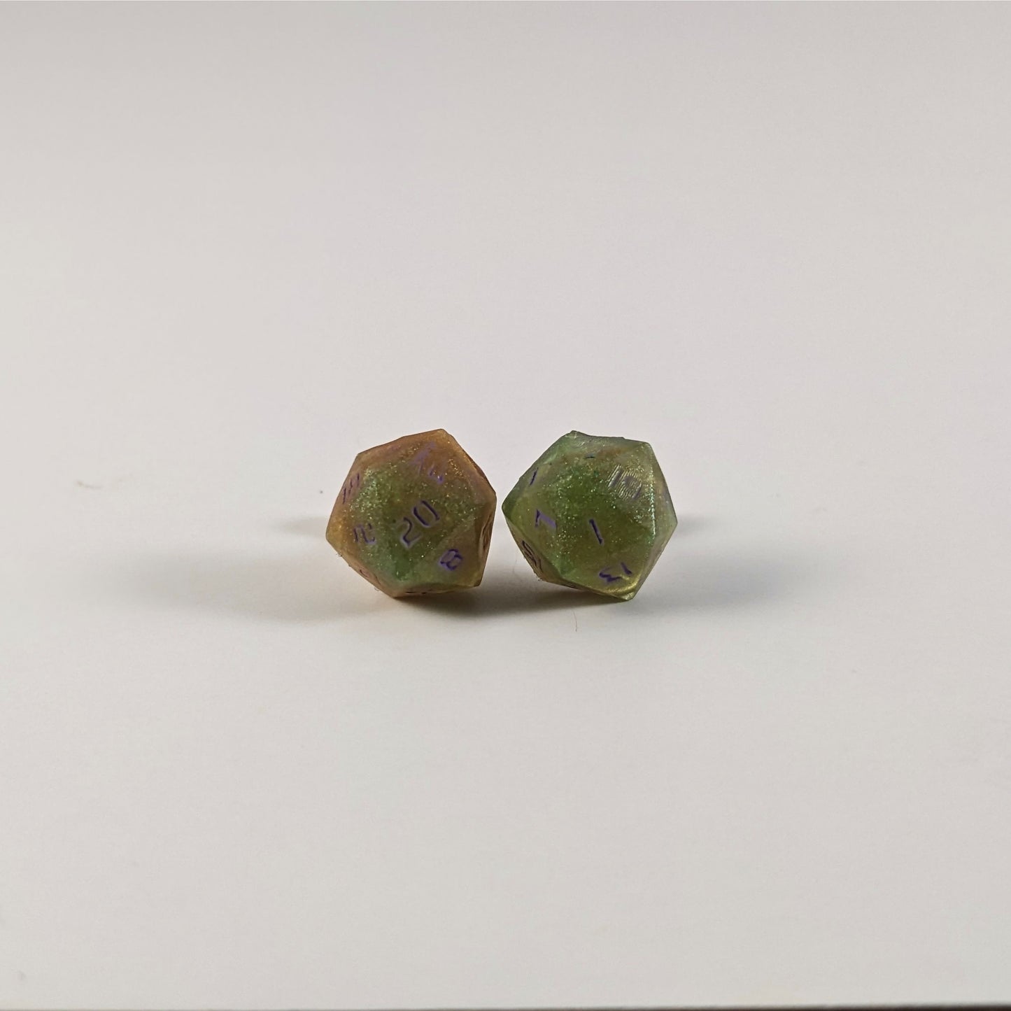 A single pair of the larger size handmade d20 earrings with sterling silver hardware that are bright green and red gold with purple numbers on a white background. The earring on the left centers the 20 face and the earring on the right centers the 1 face.