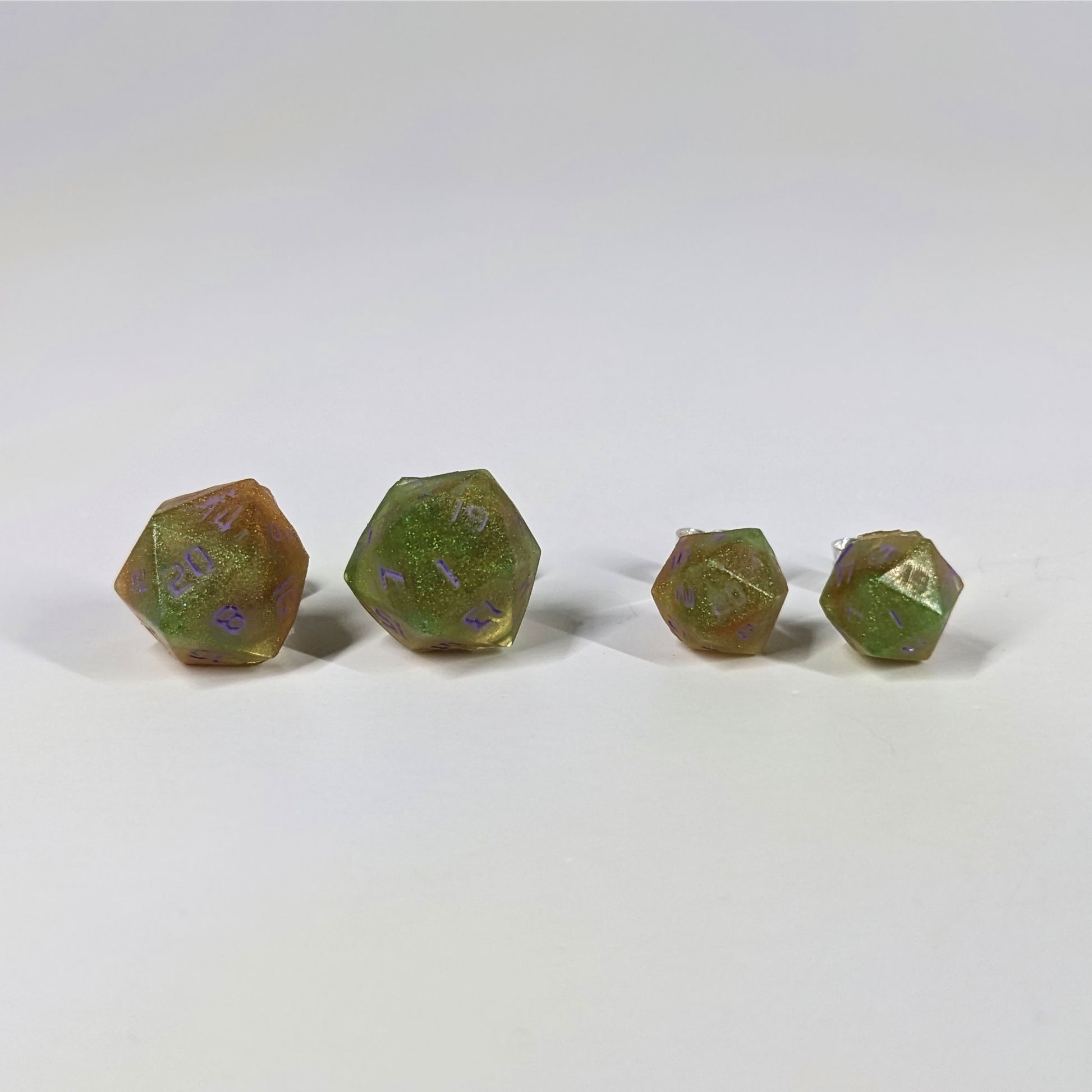 Two pairs of handmade d20 earrings with sterling silver hardware that are bright green and red gold with purple numbers on a white background sit in a line with the larger pair on the left and the smaller pair on the right. The earrings on the left of each pair centers the 20 face and the earrings on the right of each pair centers the 1 face.