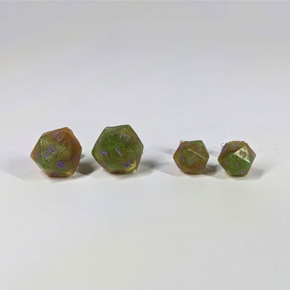 Two pairs of handmade d20 earrings with sterling silver hardware that are bright green and red gold with purple numbers on a white background sit in a line with the larger pair on the left and the smaller pair on the right. The earrings on the left of each pair centers the 20 face and the earrings on the right of each pair centers the 1 face.