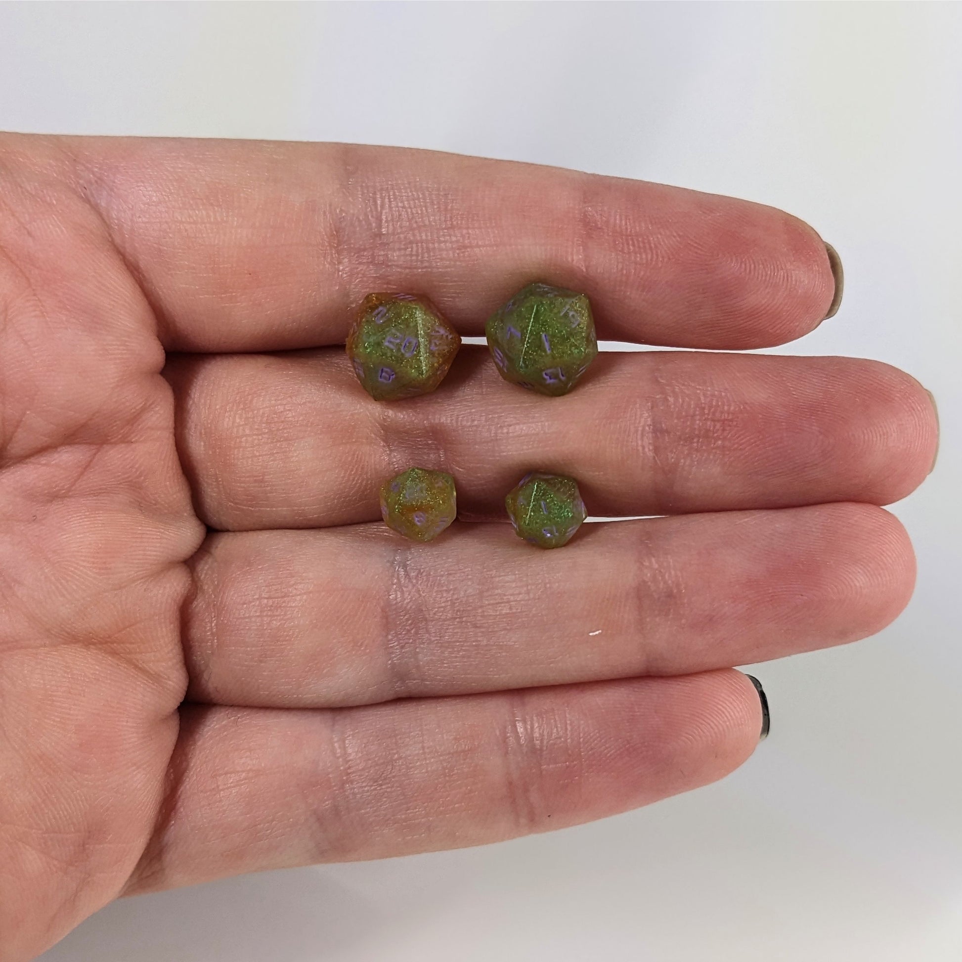 Two pairs of handmade d20 earrings with sterling silver hardware that are bright green and red gold with purple numbers held between the fingers of a flat hand. The larger pair is between the pointer and middle finger, the smaller between the middle and ring finger. The earrings on the left of each pair centers the 20 face and the earrings on the right of each pair centers the 1 face.
