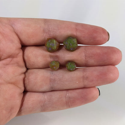 Two pairs of handmade d20 earrings with sterling silver hardware that are bright green and red gold with purple numbers held between the fingers of a flat hand. The larger pair is between the pointer and middle finger, the smaller between the middle and ring finger. The earrings on the left of each pair centers the 20 face and the earrings on the right of each pair centers the 1 face.