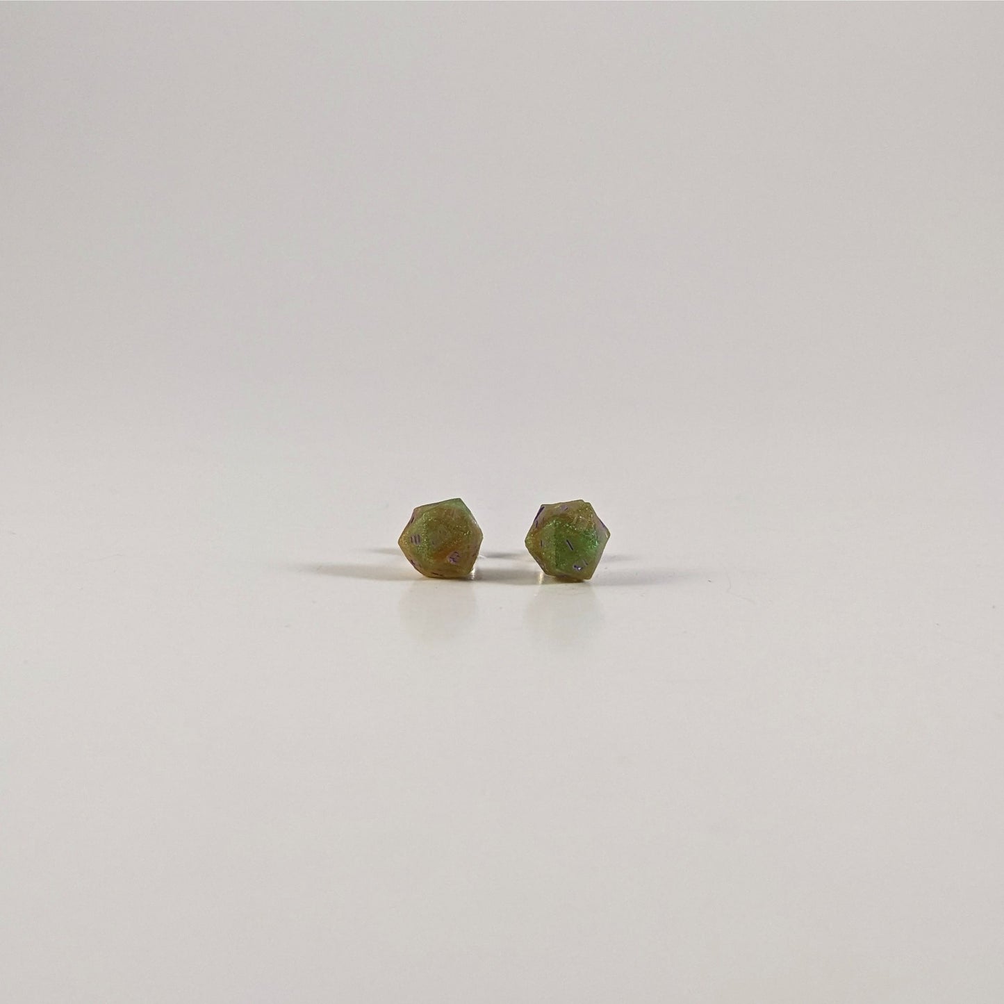 A single pair of the smaller size handmade d20 earrings with sterling silver hardware that are bright green and red gold with purple numbers on a white background. The earring on the left centers the 20 face and the earring on the right centers the 1 face.