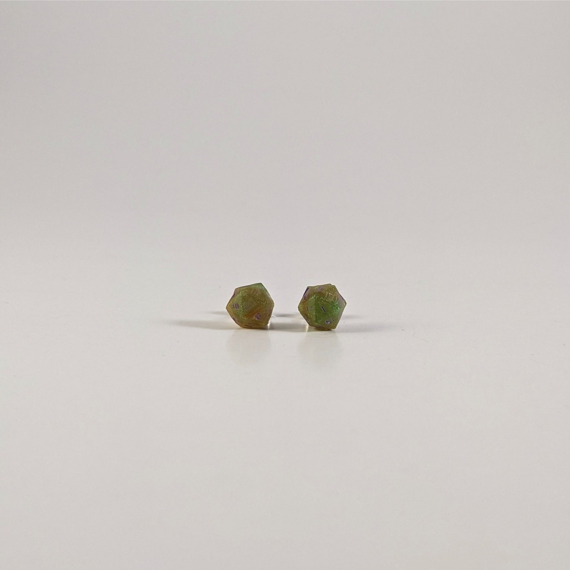 A single pair of the smaller size handmade d20 earrings with sterling silver hardware that are bright green and red gold with purple numbers on a white background. The earring on the left centers the 20 face and the earring on the right centers the 1 face.