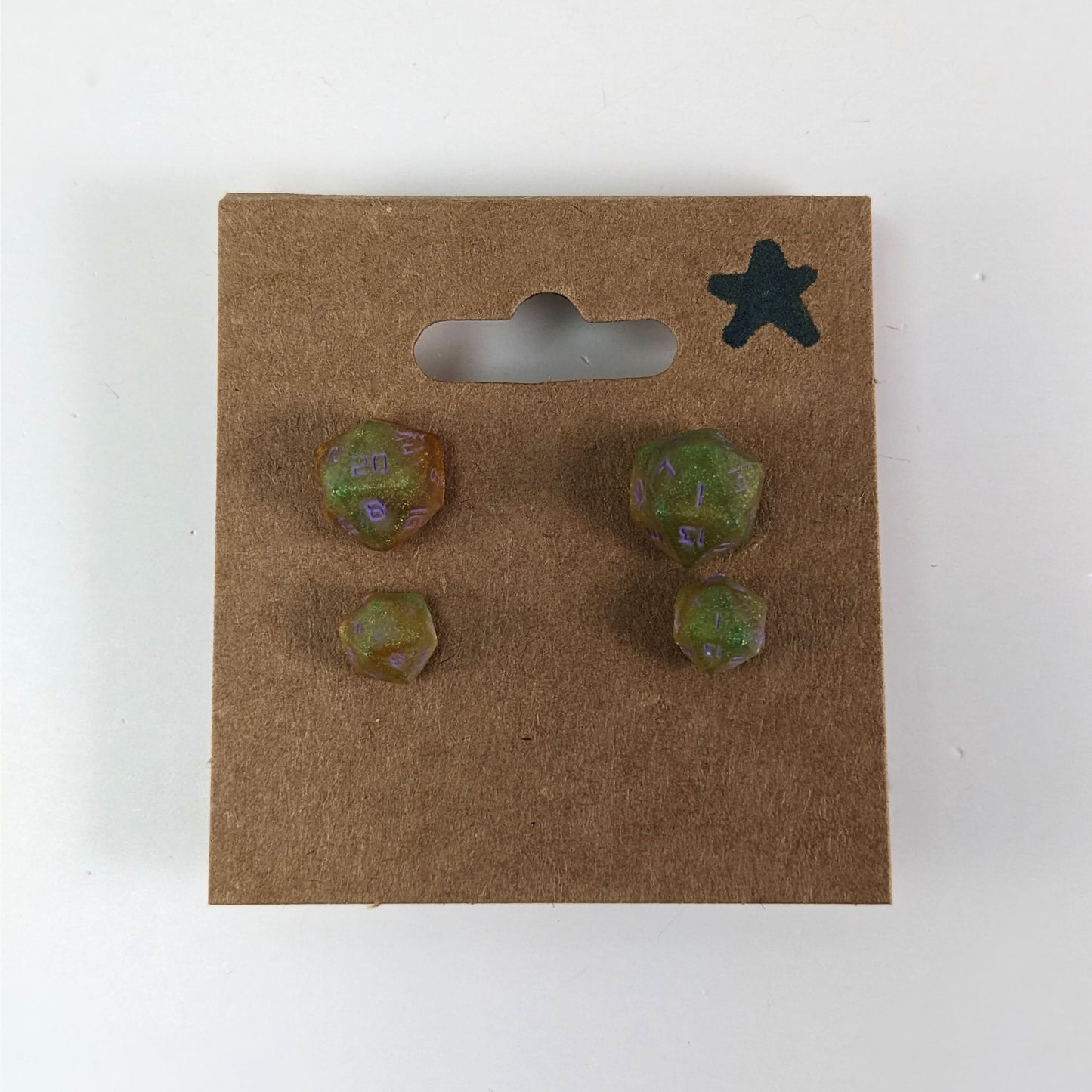 Two pairs of handmade d20 earrings with sterling silver hardware that are bright green and red gold with purple numbers on a kraft brown earring card with a star in the top right corner. The earrings on the left of each pair centers the 20 face and the earrings on the right of each pair centers the 1 face.