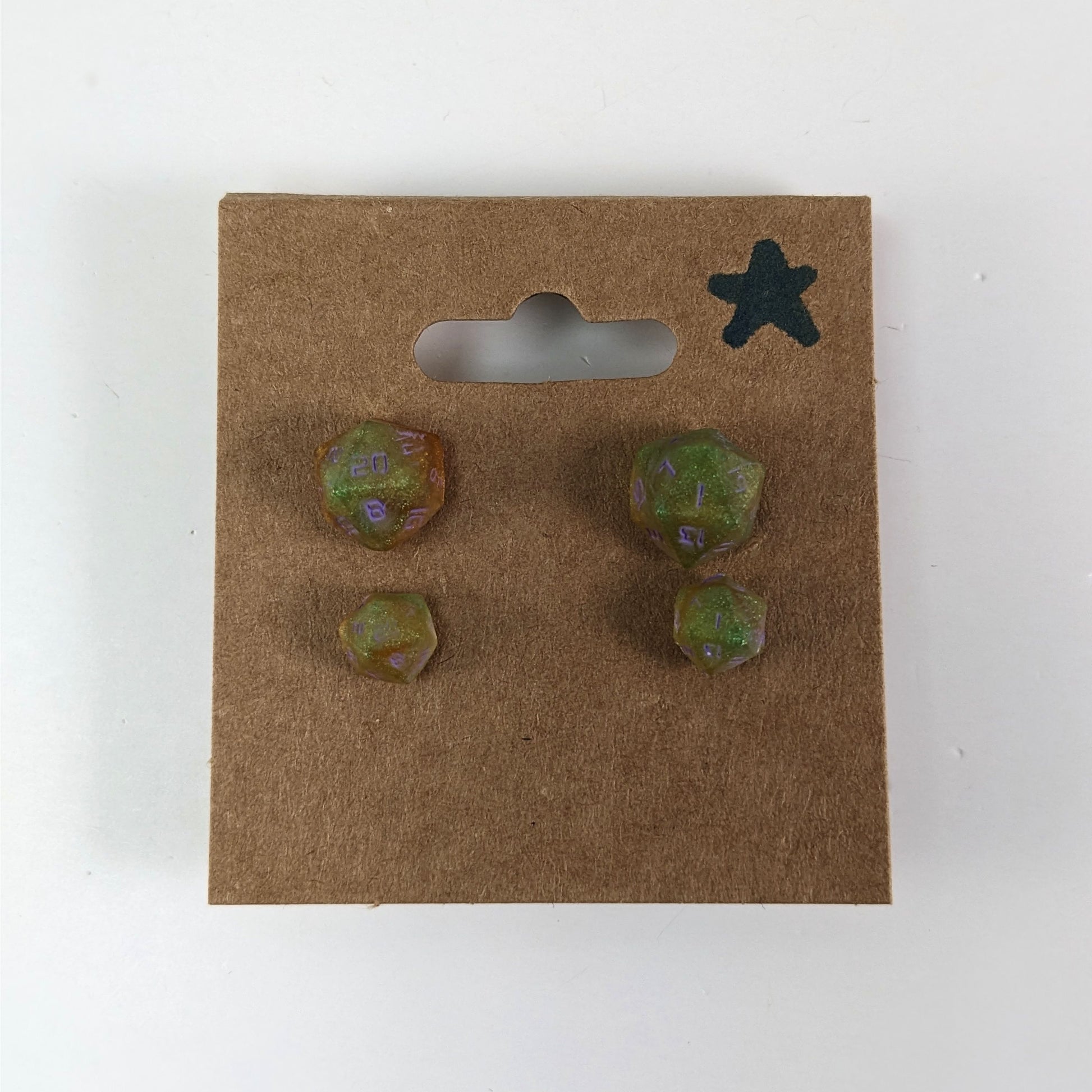 Two pairs of handmade d20 earrings with sterling silver hardware that are bright green and red gold with purple numbers on a kraft brown earring card with a star in the top right corner. The earrings on the left of each pair centers the 20 face and the earrings on the right of each pair centers the 1 face.