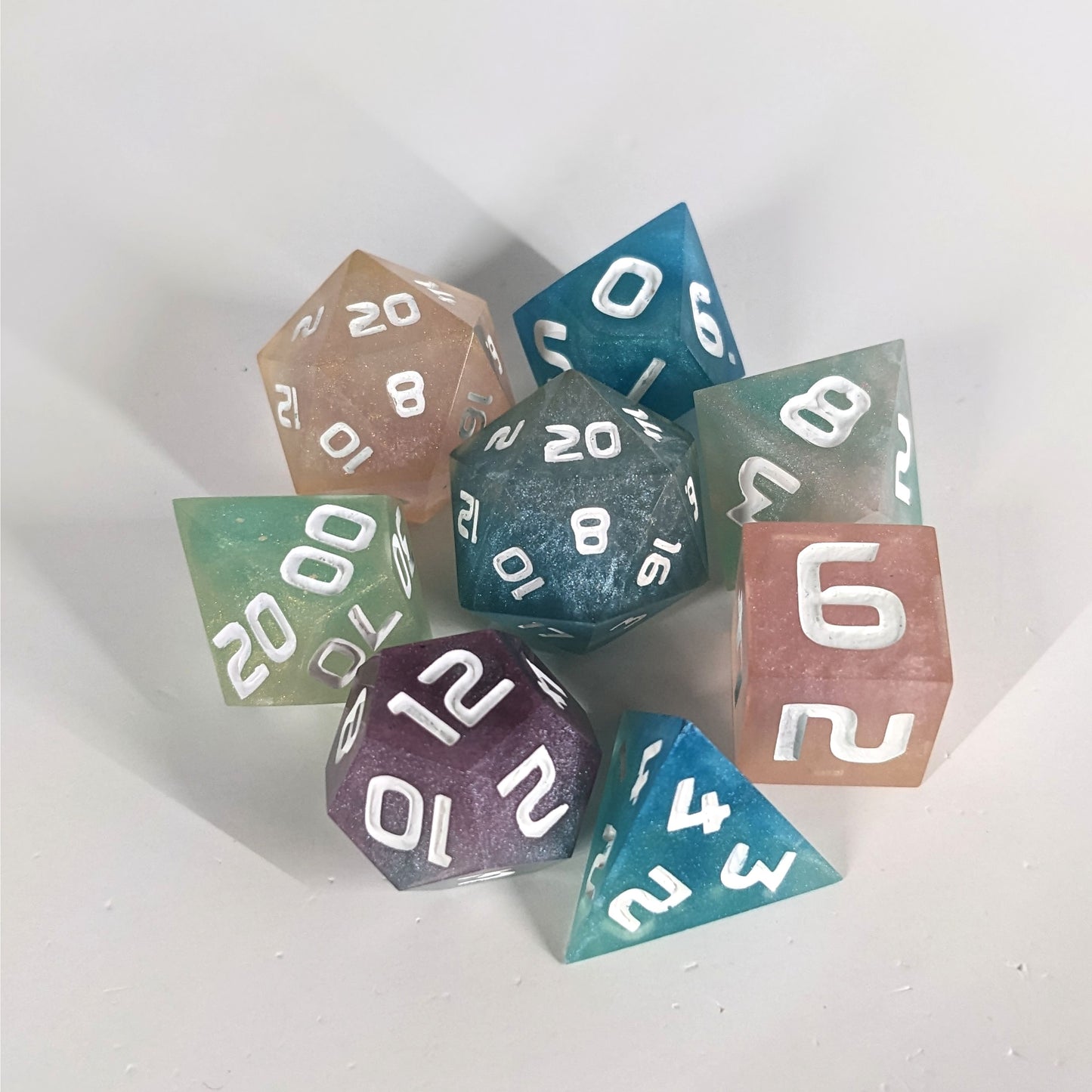 A complete set of 8 handmade artisan dice that have swirls of translucent pink, purple, green, blue, and white with opaque white numbers. The DnD dice set is arranged in a circle and features 2 D20s, one of which is in the center surrounded by the other 7 dice on a white background.