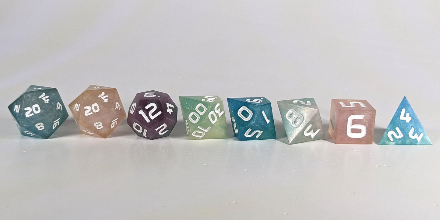 A straight on photo of a complete set of polyhedral dice that have swirls of translucent pink, purple, green, blue, and white with opaque white numbers arranged in a straight line. They are lined up with the 2 d20s on the left going down to the d4 on the right side. The highest value number of each die is facing forward