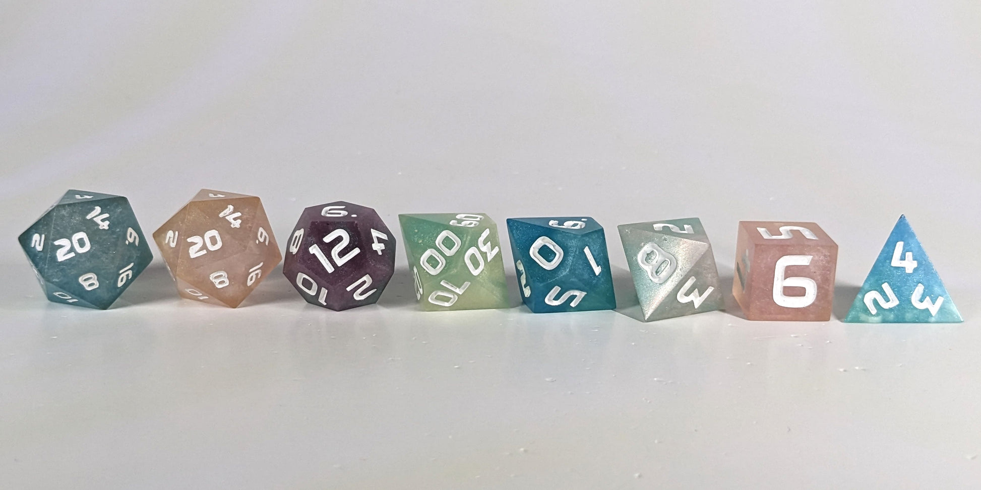 A straight on photo of a complete set of polyhedral dice that have swirls of translucent pink, purple, green, blue, and white with opaque white numbers arranged in a straight line. They are lined up with the 2 d20s on the left going down to the d4 on the right side. The highest value number of each die is facing forward