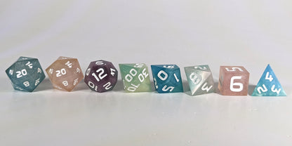 A straight on photo of a complete set of polyhedral dice that have swirls of translucent pink, purple, green, blue, and white with opaque white numbers arranged in a straight line. They are lined up with the 2 d20s on the left going down to the d4 on the right side. The highest value number of each die is facing forward