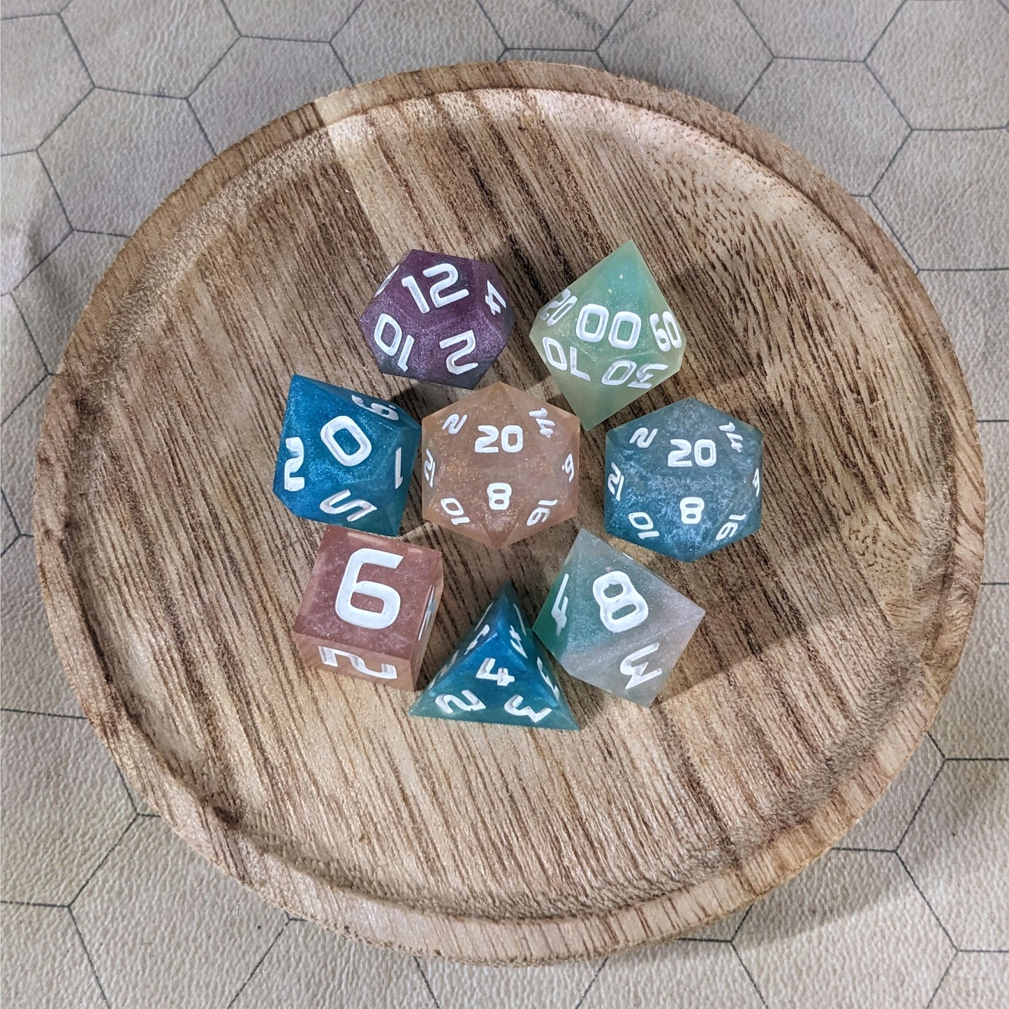 A complete set of 8 handmade artisan dice that have swirls of translucent pink, purple, green, blue, and white with opaque white numbers. The DnD dice set is on a wooded circle and the dice are arranged in a circle and features 2 D20s, one of which is in the center surrounded by the other 7 dice.