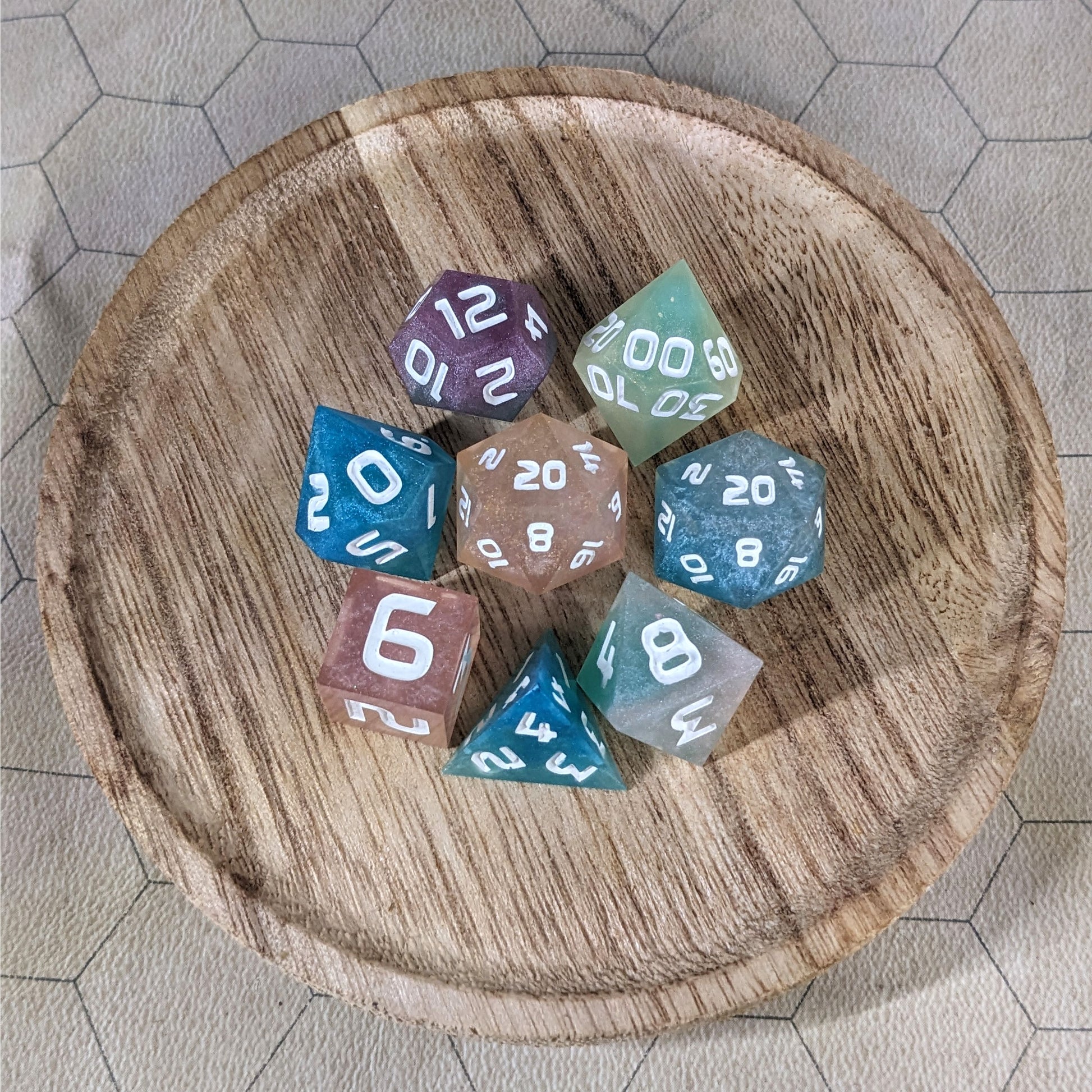 A complete set of 8 handmade artisan dice that have swirls of translucent pink, purple, green, blue, and white with opaque white numbers. The DnD dice set is on a wooded circle and the dice are arranged in a circle and features 2 D20s, one of which is in the center surrounded by the other 7 dice.