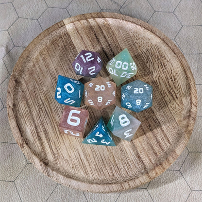 A complete set of 8 handmade artisan dice that have swirls of translucent pink, purple, green, blue, and white with opaque white numbers. The DnD dice set is on a wooded circle and the dice are arranged in a circle and features 2 D20s, one of which is in the center surrounded by the other 7 dice.
