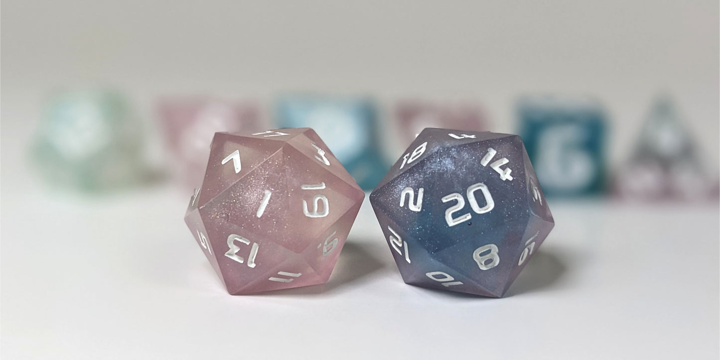 A straight on photo of a standard set of DnD dice that have swirls of translucent pink, purple, green, blue, and white with opaque white numbers focusing on 2 d20s in the center. The d20 on the left has the 1 face centered and the d20 on the right has the 20 face centered. The other 6 dice are lined up in the background but are out of focus, while the d20s are in sharp focus.