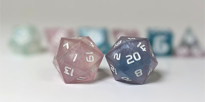 A straight on photo of a standard set of DnD dice that have swirls of translucent pink, purple, green, blue, and white with opaque white numbers focusing on 2 d20s in the center. The d20 on the left has the 1 face centered and the d20 on the right has the 20 face centered. The other 6 dice are lined up in the background but are out of focus, while the d20s are in sharp focus.