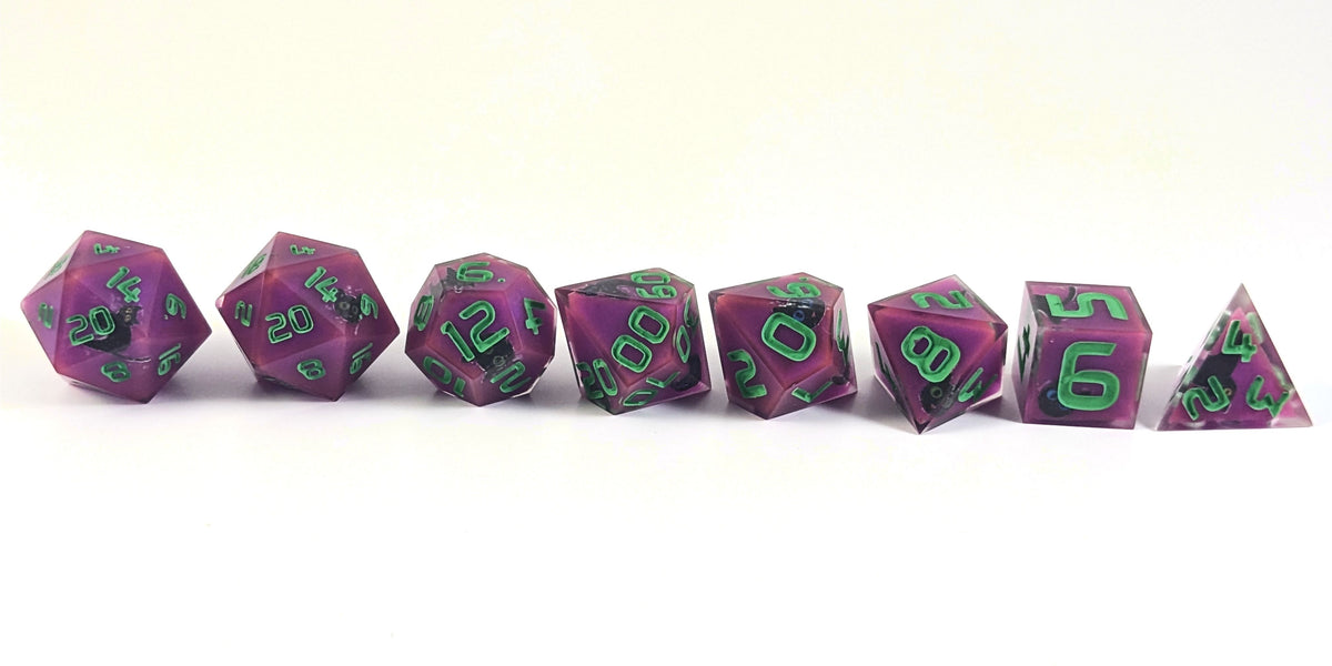 A straight on photo of a complete set of polyhedral dice that are purple with black cat designs with bright green numbers arranged in a straight line. They are lined up with the 2 d20s on the left going down to the d4 on the right side. The highest value number of each die is facing forward