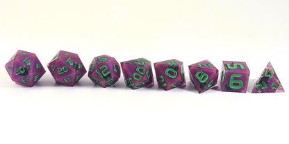 A straight on photo of a complete set of polyhedral dice that are purple with black cat designs with bright green numbers arranged in a straight line. They are lined up with the 2 d20s on the left going down to the d4 on the right side. The highest value number of each die is facing forward