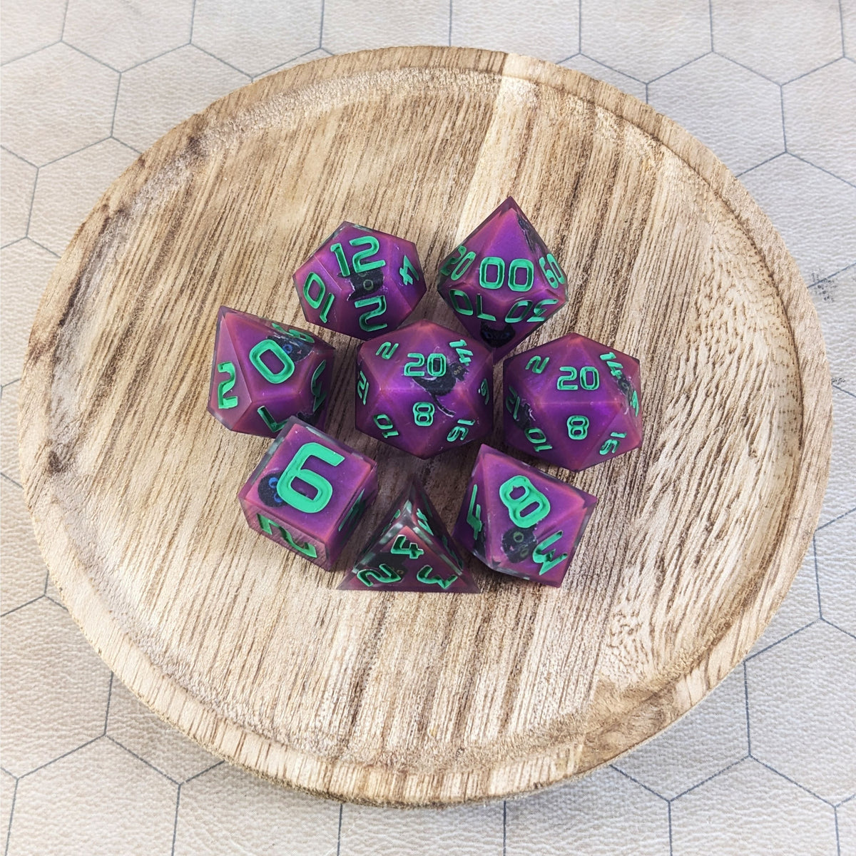 A complete set of 8 handmade artisan dice that are purple with black cat designs with bright green numbers. The DnD dice set is on a wooded circle and the dice are arranged in a circle and features 2 D20s, one of which is in the center surrounded by the other 7 dice.