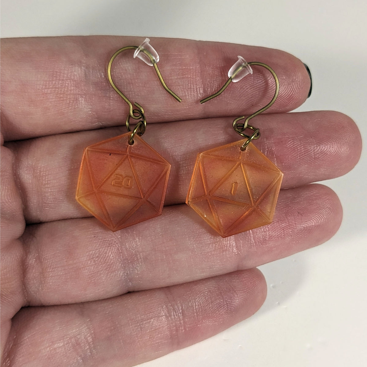 A pair of fiery red and orange dangly, flat d20 earrings with gold color titanium and niobium hardware with bell shaped rubber backs held on on a hand on a white background. The earring on the left has a 20 on it and the earring on the right has a 1 on it, both in a sci-fi style font.