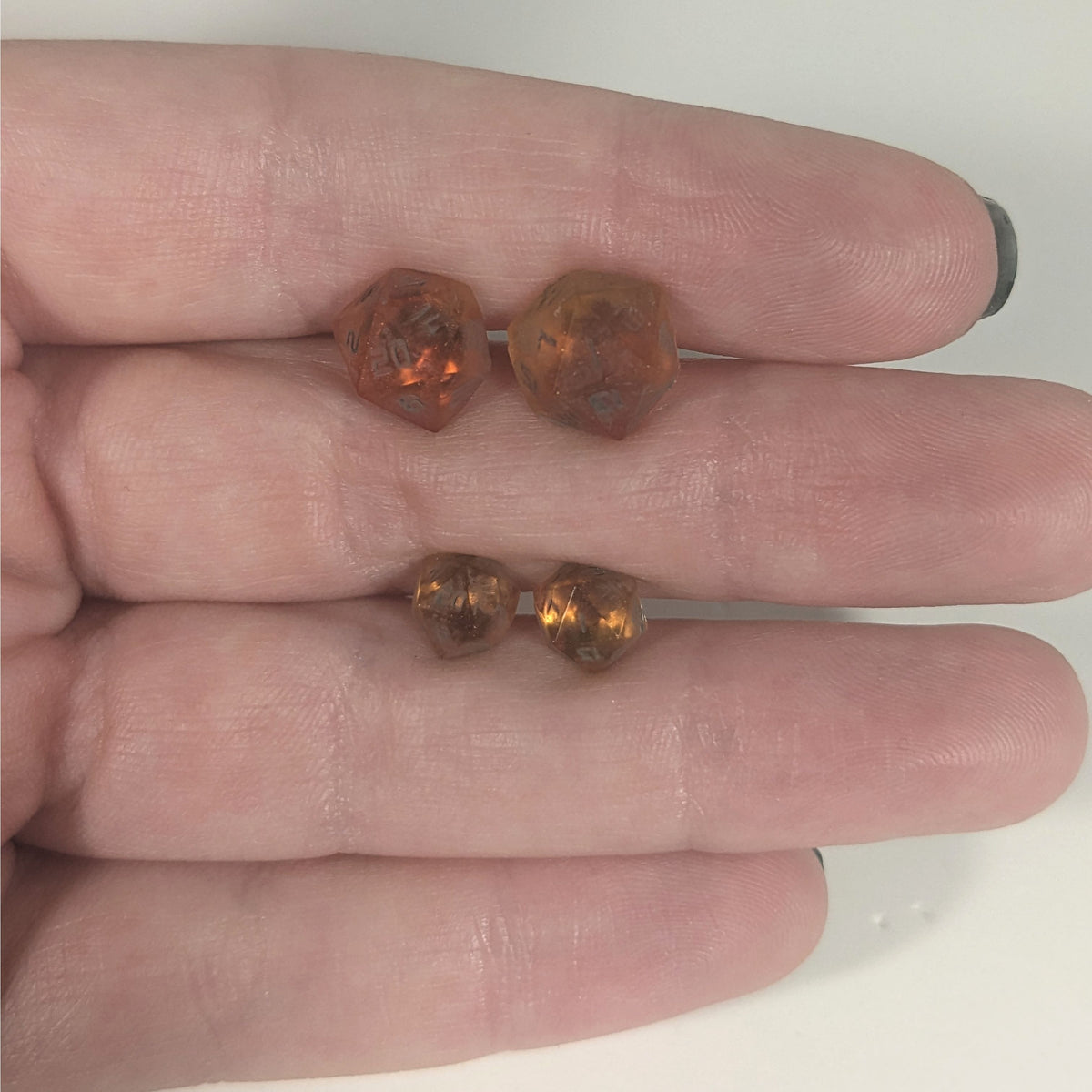 Two pairs of handmade d20 earrings with titanium hardware that are red and orange swirls with smokey gray numbers held between the fingers of a flat hand. The larger pair is between the pointer and middle finger, the smaller between the middle and ring finger. The earrings on the left of each pair centers the 20 face and the earrings on the right of each pair centers the 1 face.
