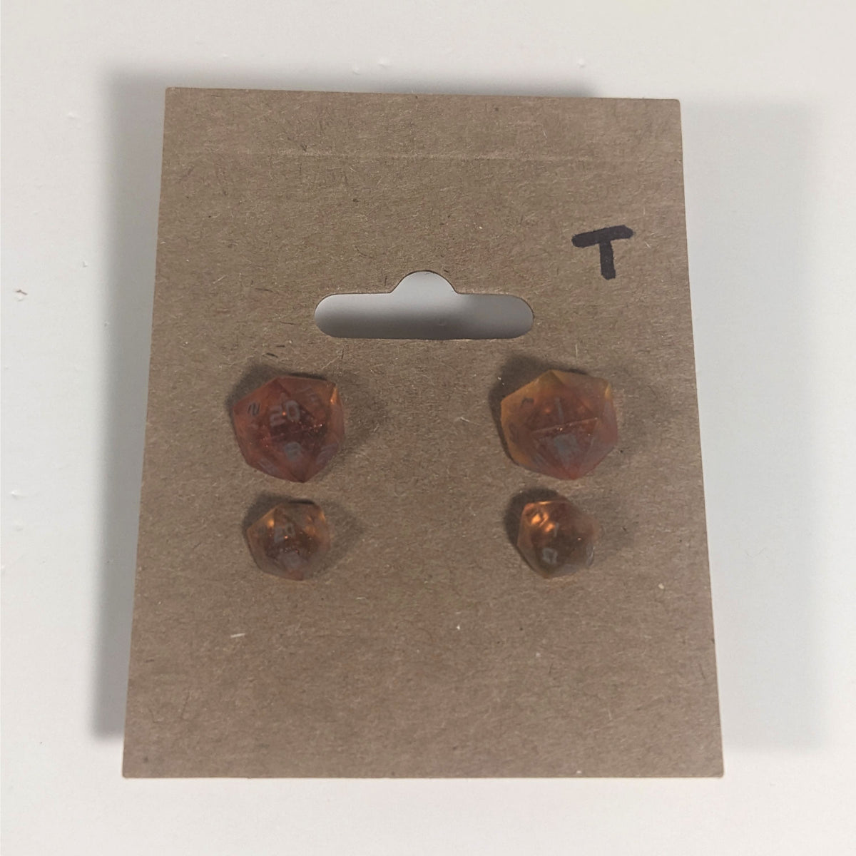 Two pairs of handmade d20 earrings with titanium hardware that are red and orange swirls with smokey gray numbers on a kraft brown earring card with a T in the top right corner. The earrings on the left of each pair centers the 20 face and the earrings on the right of each pair centers the 1 face.