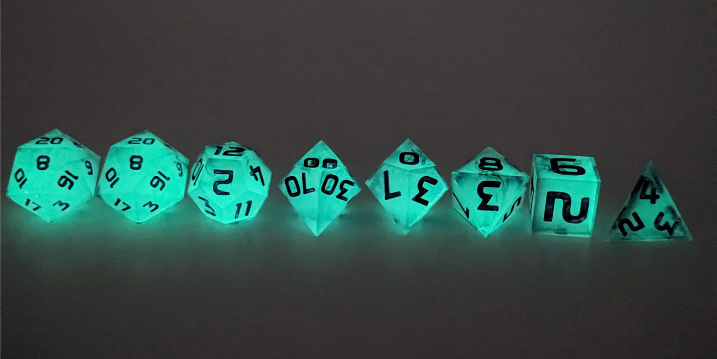A straight on photo of a complete set of polyhedral dice that have glowing green inserts and have purple numbers arranged in a straight line. They are lined up with the 2 d20s on the left going down to the d4 on the right side. The highest value number of each die is facing up. This image shows the glow-in-the-dark feature of this dice set and that you can still see the black swirls through the glow.