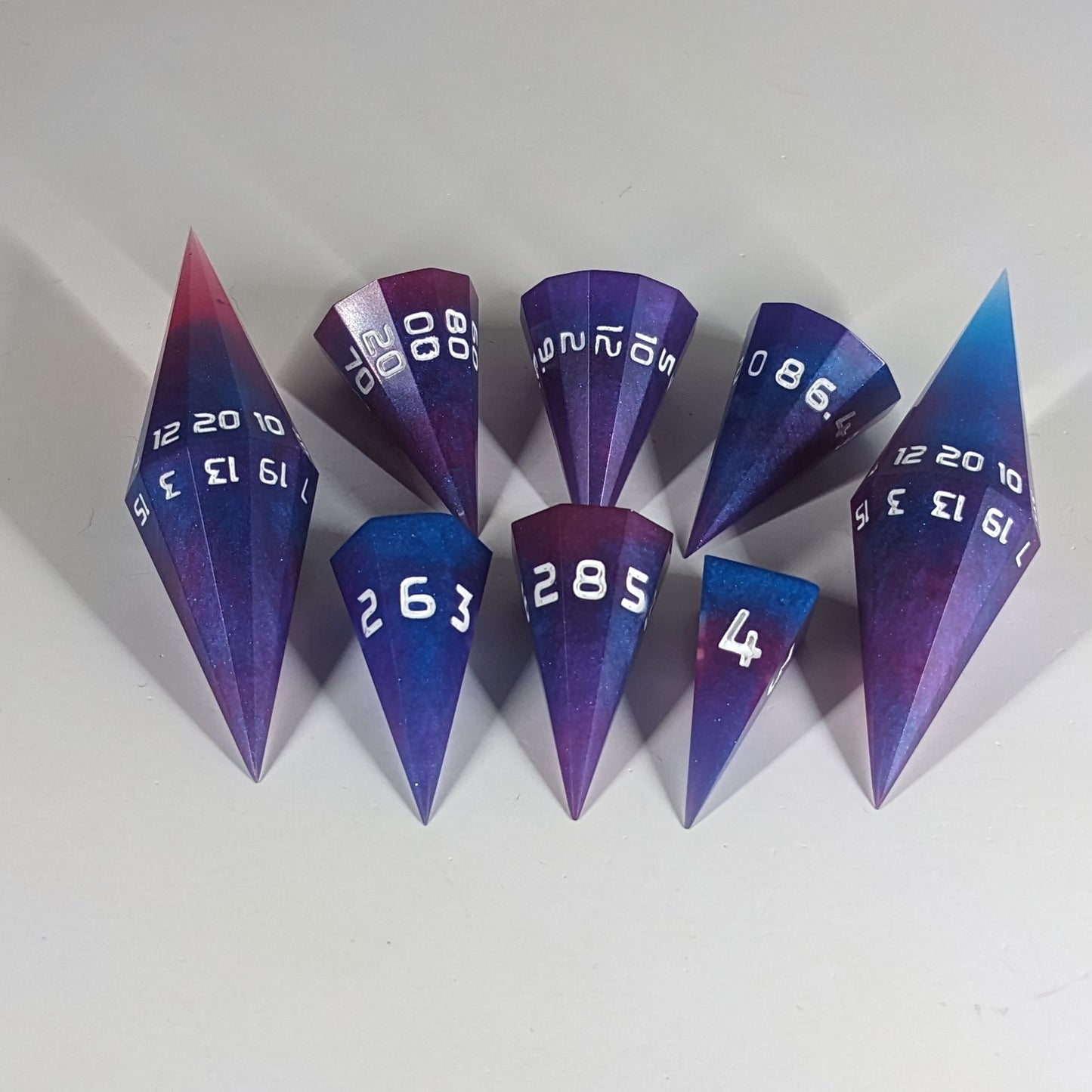 The dagger style DnD dice set that is purple, pink, and blue galaxy swirls with an extra touch of glitter and white numbers is arranged with a d20 each on the right and left, with the d%, d12, and d10 along the top and the d6, d8, and d4 along the bottom, all in line with the d20.