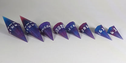 A straight on photo of a complete set of dagger DnD dice that are purple, pink, and blue galaxy swirls with an extra touch of glitter and white numbers arranged in a straight line with points angled toward the right. They are lined up with the 2 d20s on the left going down to the d4 on the right side. The highest value number of each die is facing forward.