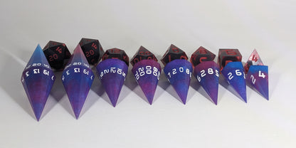 A straight on photo of a complete set of dagger dice that are purple, pink, and blue galaxy swirls with an extra touch of glitter and white numbers arranged in a straight line with points angled forward. They are lined up with the 2 d20s on the left going down to the d4 on the right side. Behind the dagger set is a standard set of polyhedral dice to allow for size comparison. The dagger dice are much larger overall, but not taller. The highest value number of each die is facing up.