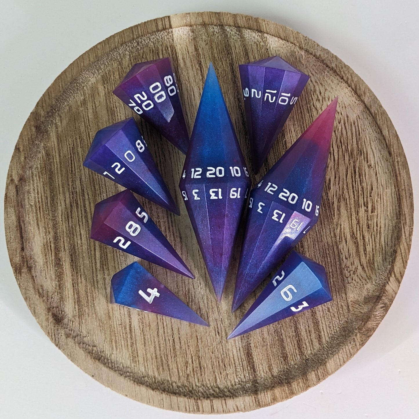 A complete set of purple, pink, and blue galaxy swirls with an extra touch of glitter and white numbers dagger style resin dice are arranged on a wooden circle. The 2 d20s are next to each other with the d12 placed between them on top. The d6 is to the bottom right of the d20s and the rest of the dice are arranged along the left side, points angled toward the d20s.