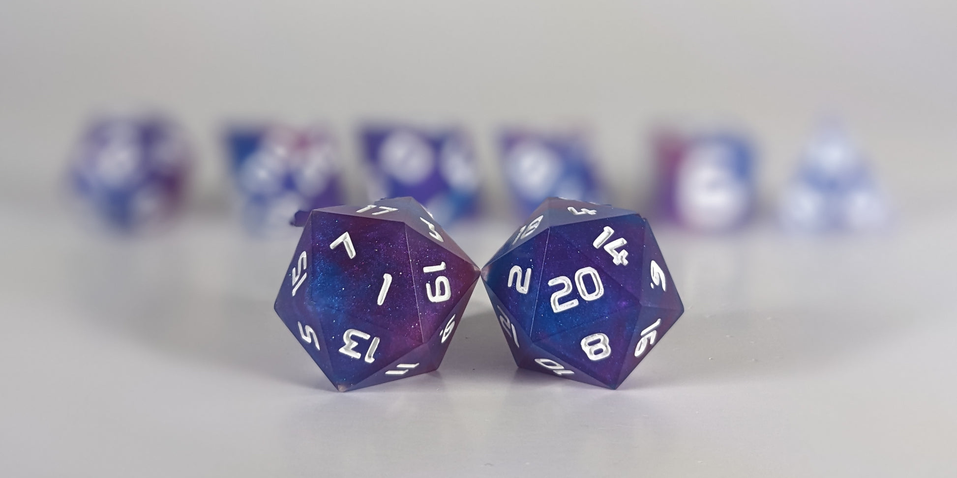 A straight on photo of a standard set of dice that are purple, pink, and blue swirls with a clear shell with extra glitter and white numbers focusing on 2 d20s in the center. The d20 on the left has the 1 face centered and the d20 on the right has the 20 face centered. The other 6 dice are lined up in the background but are out of focus, while the d20s are in sharp focus.