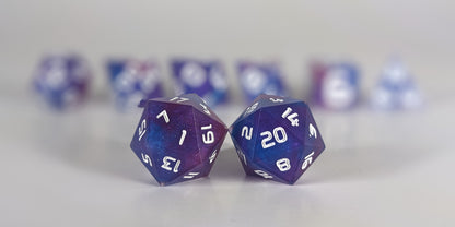 A straight on photo of a standard set of dice that are purple, pink, and blue swirls with a clear shell with extra glitter and white numbers focusing on 2 d20s in the center. The d20 on the left has the 1 face centered and the d20 on the right has the 20 face centered. The other 6 dice are lined up in the background but are out of focus, while the d20s are in sharp focus.