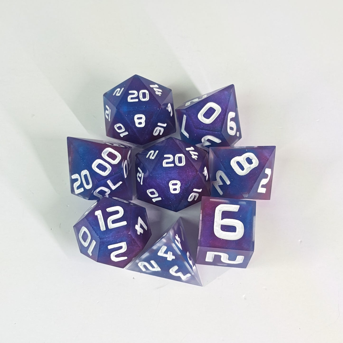 A complete set of 8 handmade artisan dice that are purple, pink, and blue swirls with a clear shell with extra glitter and white numbers. The DnD dice set is arranged in a circle and features 2 D20s, one of which is in the center surrounded by the other 7 dice.