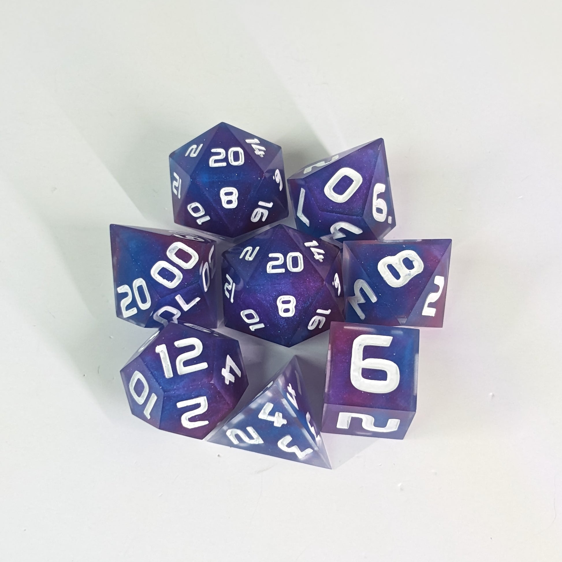 A complete set of 8 handmade artisan dice that are purple, pink, and blue swirls with a clear shell with extra glitter and white numbers. The DnD dice set is arranged in a circle and features 2 D20s, one of which is in the center surrounded by the other 7 dice.