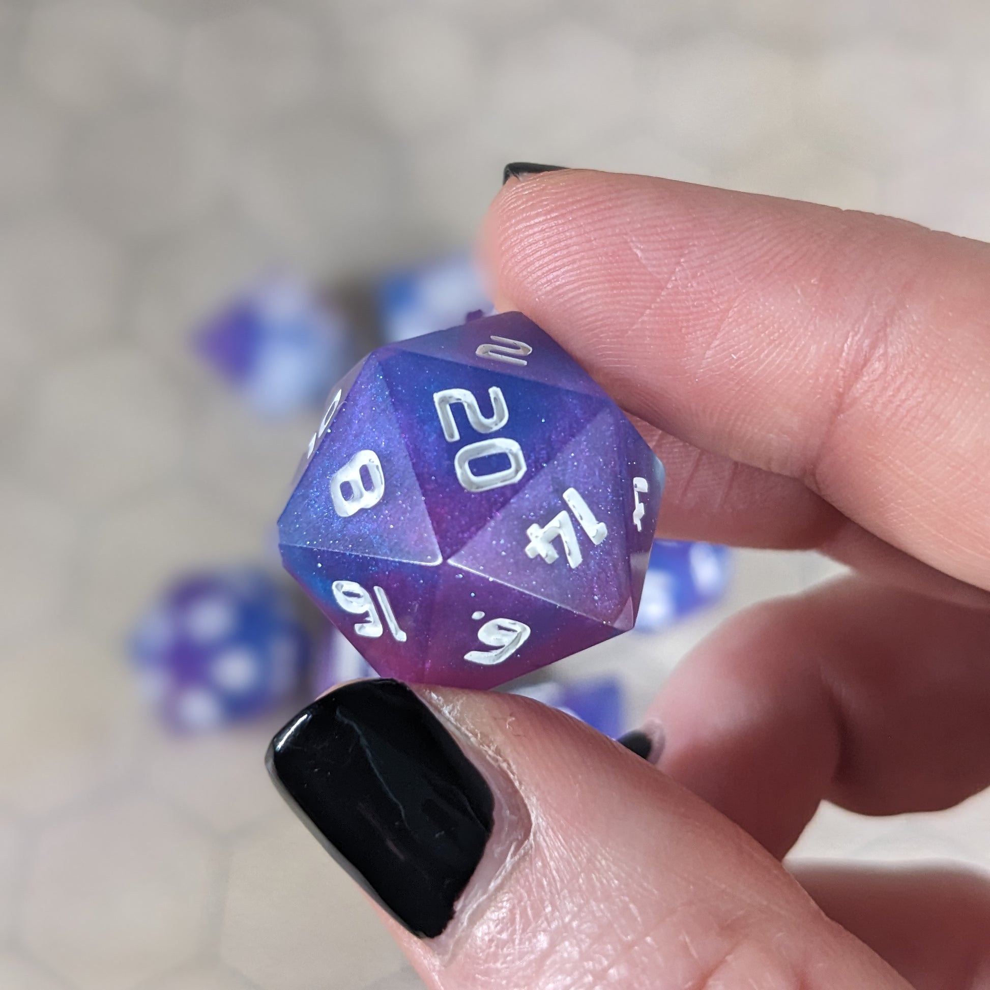 A d20 that is purple, pink, and blue swirls with a clear shell with extra glitter and white numbers is held up to the camera to show off the color swirls and glittery shell. The 20 face is angled toward the camera.