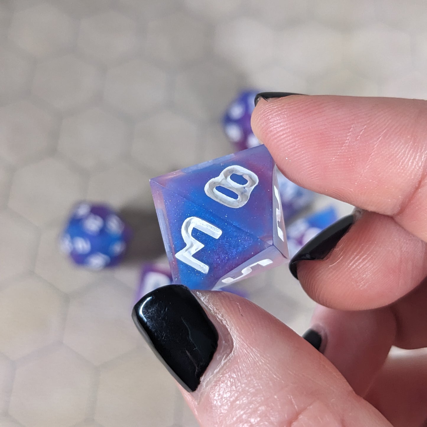 A d8 that is purple, pink, and blue swirls with a clear shell with extra glitter and white numbers is held up to the camera to show off the color swirls and glittery shell. The 8 and 3 faces are angled toward the camera.