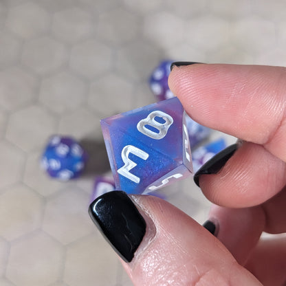 A d8 that is purple, pink, and blue swirls with a clear shell with extra glitter and white numbers is held up to the camera to show off the color swirls and glittery shell. The 8 and 3 faces are angled toward the camera.