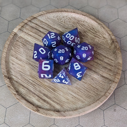 A complete set of 8 handmade artisan dice that are purple, pink, and blue swirls with a clear shell with extra glitter and white numbers. The DnD dice set is on a wooded circle and the dice are arranged in a circle and features 2 D20s, one of which is in the center surrounded by the other 7 dice.