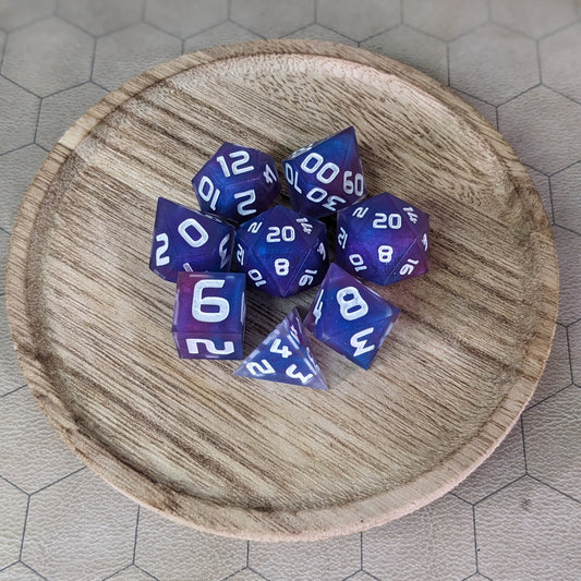 A complete set of 8 handmade artisan dice that are purple, pink, and blue swirls with a clear shell with extra glitter and white numbers. The DnD dice set is on a wooded circle and the dice are arranged in a circle and features 2 D20s, one of which is in the center surrounded by the other 7 dice.