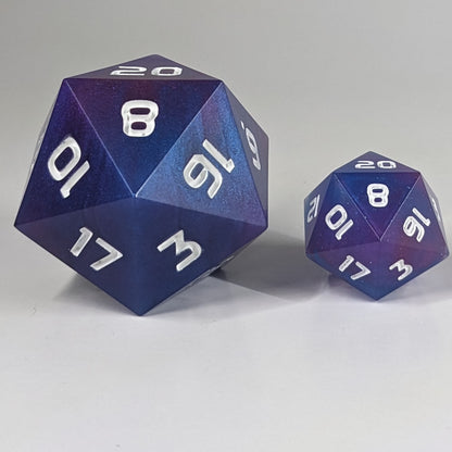 A straight on shot of a handcrafted large d20 that has subtle swirls of purple, blue, and pink with white numbers to the left of a standard size d20 that also has swirls of blue, pink, and purple, but also more sparkle. This size comparison shows the large d20 being about twice the size of the standard.