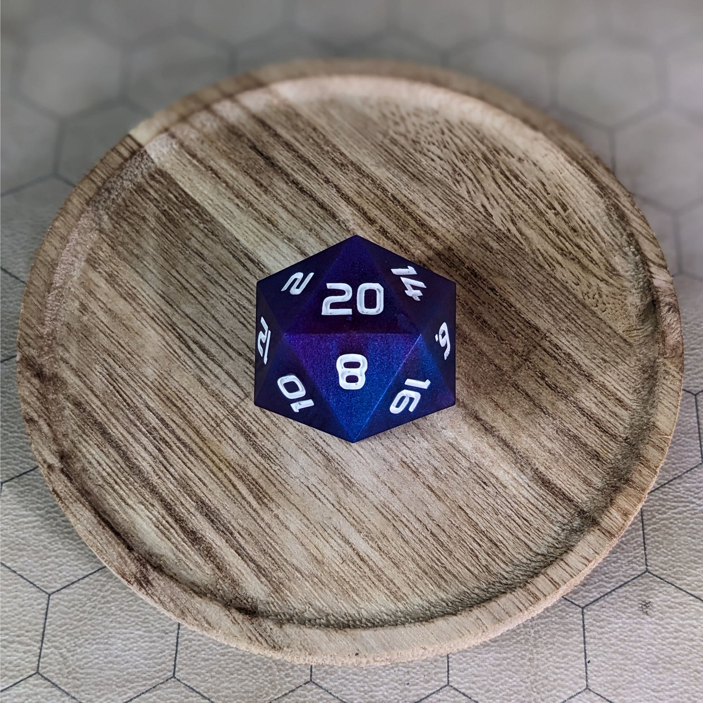 A high angle view of a large d20 with subtle swirls of purple, blue, and pink with white numbers. It is sitting in the center of a wooden circle.