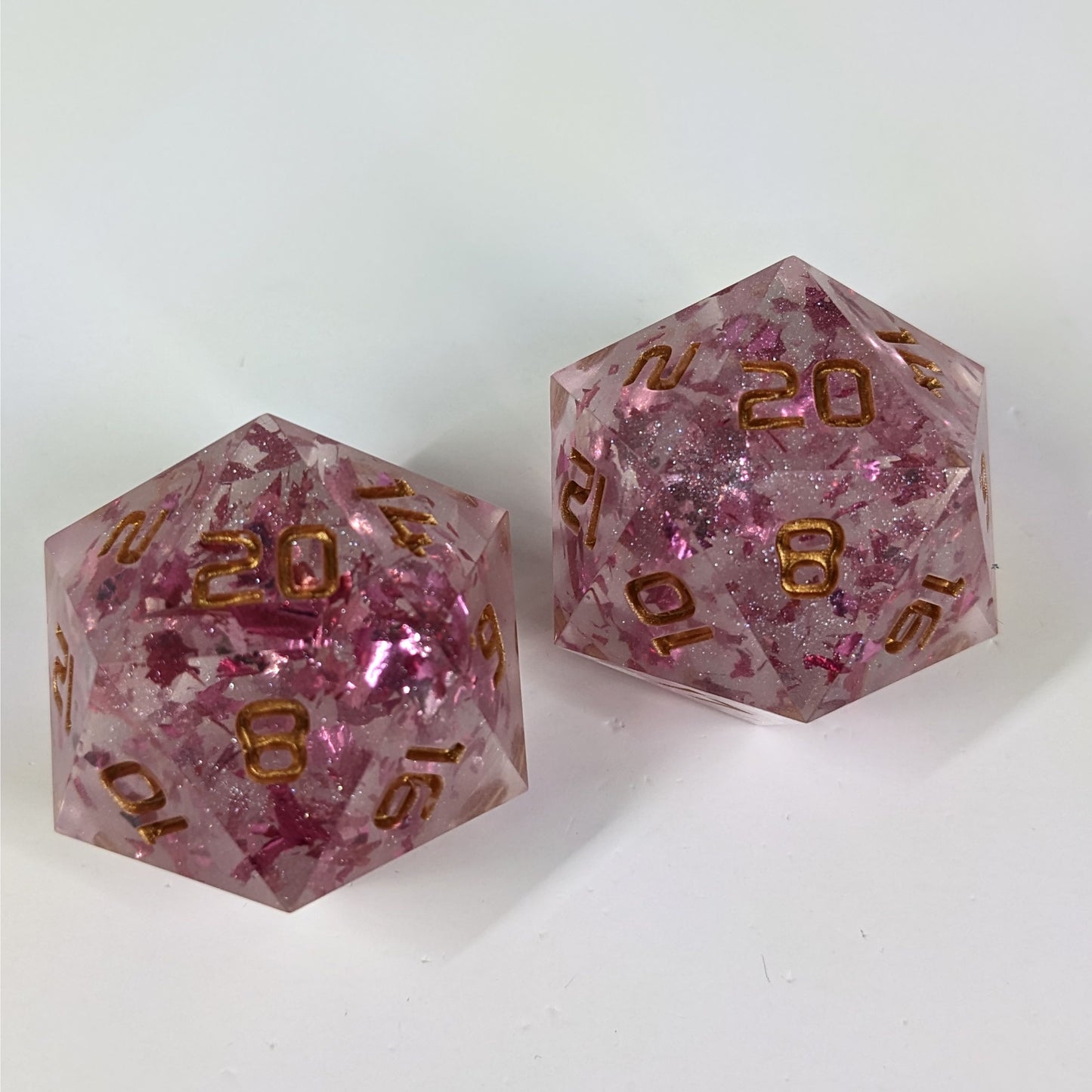A high angle photo of two large d20 that are clear with metallic pink flakies and glitter and gold numbers on a white background.