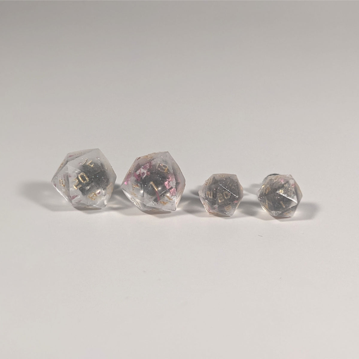 Two pairs of handmade d20 earrings with titanium hardware that are glittery clear with pink flakies with gold numbers on a white background sit in a line with the larger pair on the left and the smaller pair on the right. The earrings on the left of each pair centers the 20 face and the earrings on the right of each pair centers the 1 face.