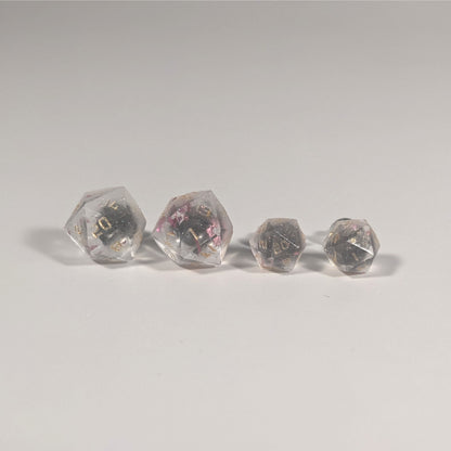 Two pairs of handmade d20 earrings with titanium hardware that are glittery clear with pink flakies with gold numbers on a white background sit in a line with the larger pair on the left and the smaller pair on the right. The earrings on the left of each pair centers the 20 face and the earrings on the right of each pair centers the 1 face.