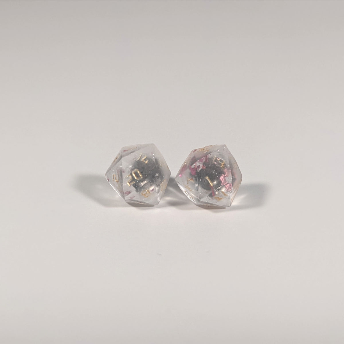 A single pair of the larger size handmade d20 earrings with titanium hardware that are glittery clear with pink flakies with gold numbers on a white background. The earring on the left centers the 20 face and the earring on the right centers the 1 face.