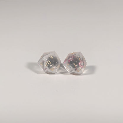 A single pair of the larger size handmade d20 earrings with titanium hardware that are glittery clear with pink flakies with gold numbers on a white background. The earring on the left centers the 20 face and the earring on the right centers the 1 face.