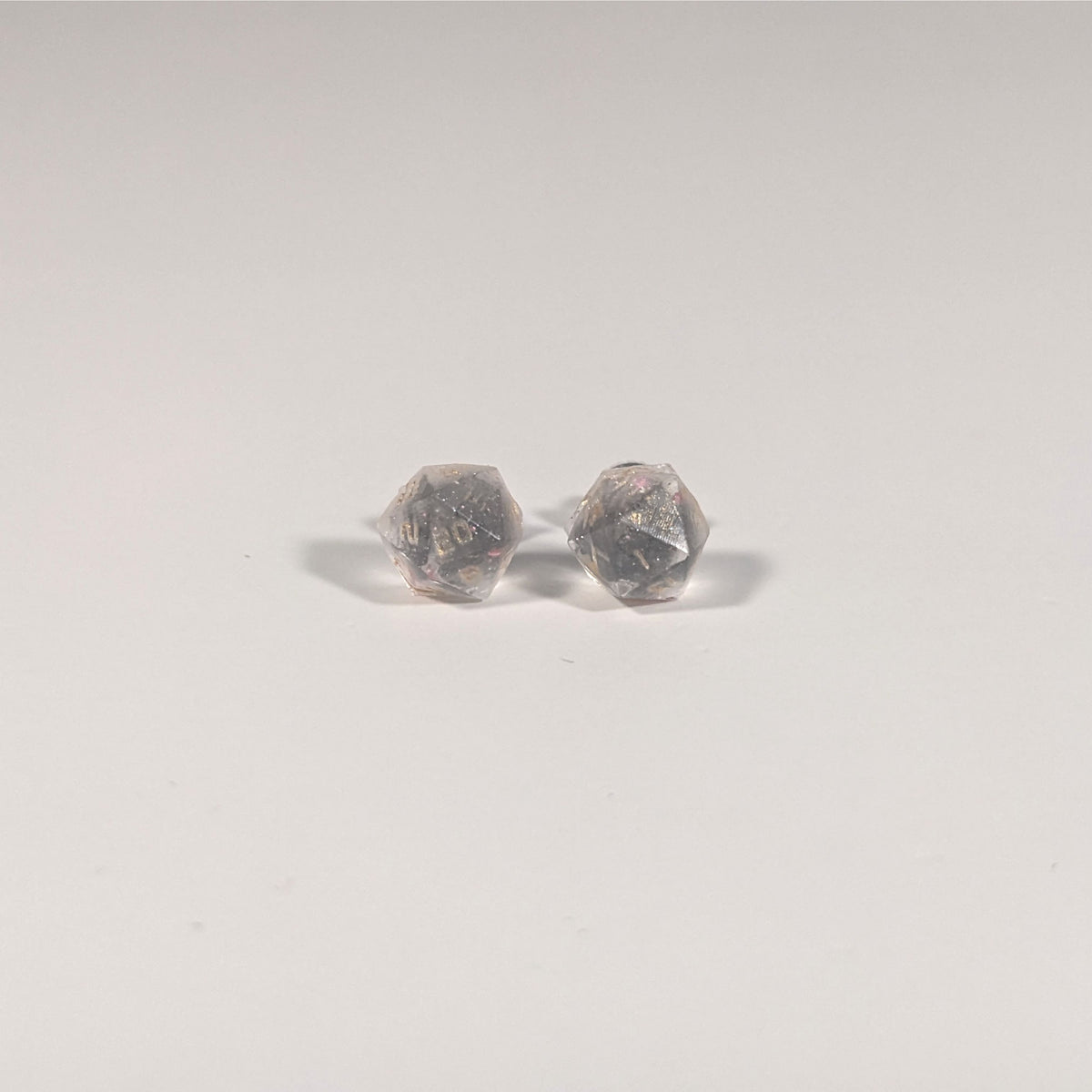 A single pair of the smaller size handmade d20 earrings with titanium hardware that are glittery clear with pink flakies with gold numbers on a white background. The earring on the left centers the 20 face and the earring on the right centers the 1 face.